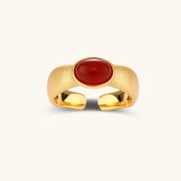 Close-up of brushed gold-plated ring with deep red oval stone, featuring adjustable band design on neutral background.